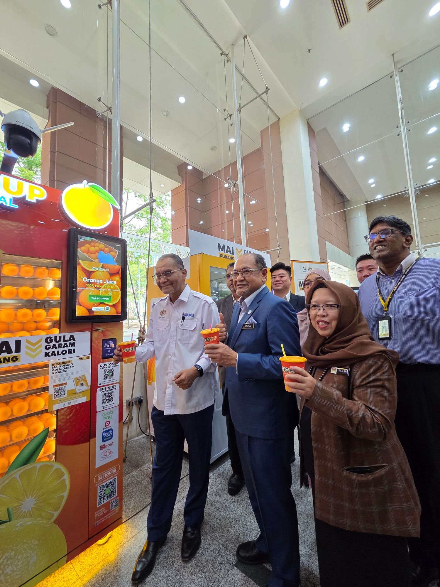 Ministry officials holding Juicesup orange juice cups beside the vending machine